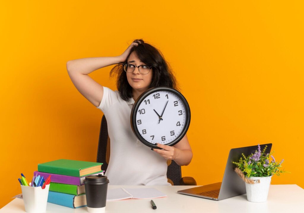Stressed student girl holding a clock and placing a hand on her head while sitting at a study desk against an orange background.