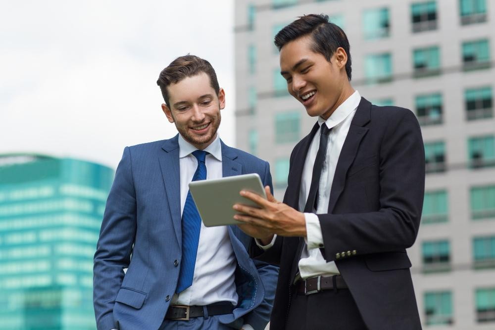 Two businessmen smiling while discussing something on a tablet in an outdoor office environment.