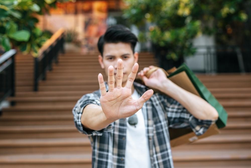 Young man holding shopping bags while showing a stop gesture with his hand in front of stairs.