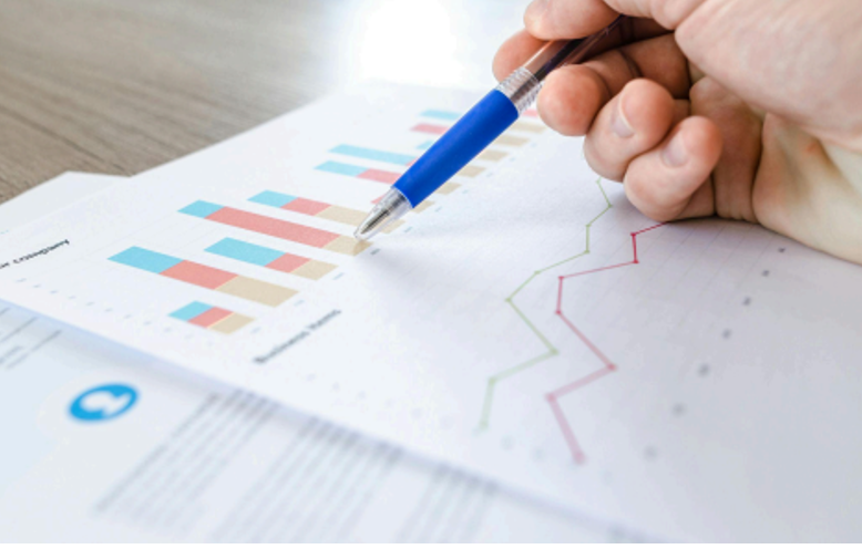 : Close-up of a hand pointing to colorful business performance charts and data graphs with a pen.
