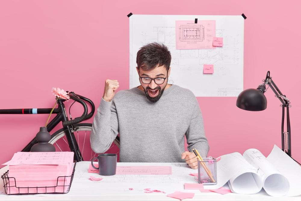Excited man celebrating success at a creative desk setup with pink decor and architectural plans.