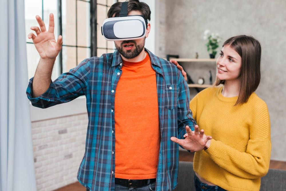 A man wearing a VR headset with his hand extended, appearing to interact with virtual elements, while a woman in a yellow sweater watches beside him. This represents virtual reality technology, immersive experiences, and emerging tech adoption.
