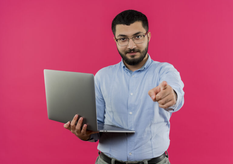 Confident man with laptop pointing at the camera against a pink background.